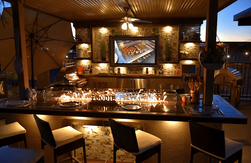 At dusk, the Kokomo Grills Outdoor Kitchen Montego Model BBQ Island and BBQ Grill sits under a wooden pergola, with cushioned bar stools, string lights on the counter, cooking utensils, and a large TV overhead.