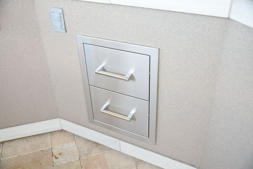 The Kokomo Grills Outdoor Kitchen Stainless Steel Double Drawer KO-DD12 by Kokomo Grills is built into a beige wall above a tiled floor, next to a light switch or outlet.