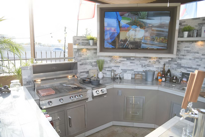 The outdoor kitchen features a Kokomo Grills KO-DD12 stainless steel double drawer, built-in grill, countertop appliances, assorted bottles, and a large TV showing a cooking show. The well-lit space has tiled walls and nearby plants.