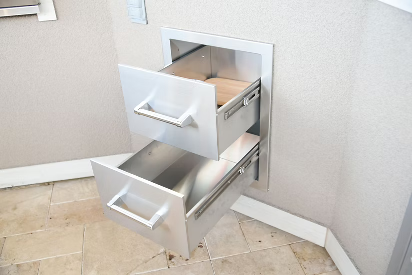 The Kokomo Grills Outdoor Kitchen Stainless Steel Double Drawer KO-DD12, by Kokomo Grills, is built into a light-colored wall. Both metal drawers are partially open, showing light wood interiors above beige tiled flooring.