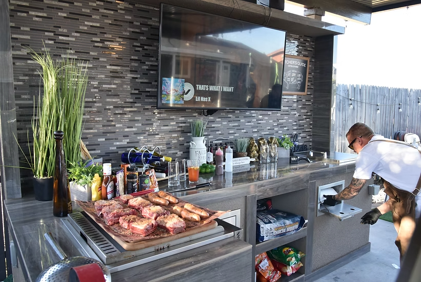 A man preps food at an outdoor kitchen with modern backsplash, TV, and a counter of seasonings, sauces, drinks, and raw steaks. A Kokomo Grills Outdoor Kitchen Stainless Steel Paper Towel Holder KO-ATH is handy on the shelves below near the snacks.