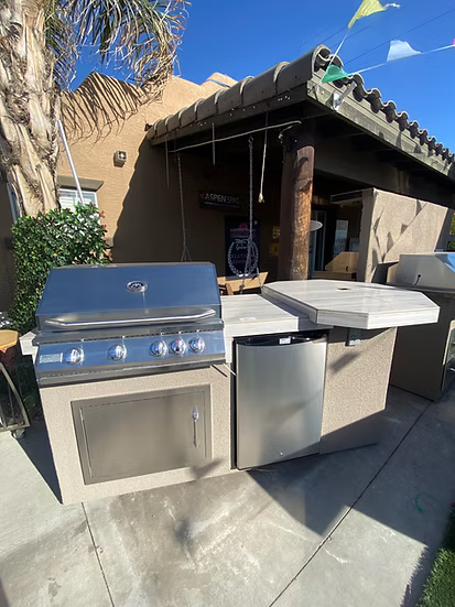 Outdoor kitchen with a Kokomo Grills Poly 7′6” High Bar BBQ Kitchen Island, closed cabinet, mini fridge, and counters under a tiled patio roof. Palm trees and a blue sky complete the scene.