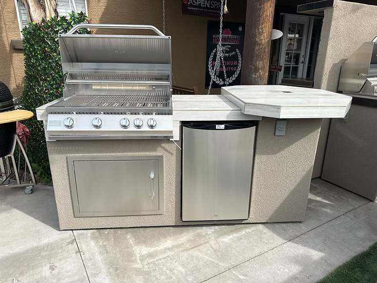 A Kokomo Grills Poly 7′6” High Bar BBQ Kitchen Island with a stainless steel 4-knob grill, built-in mini fridge, countertop with sink, all set on a concrete patio.
