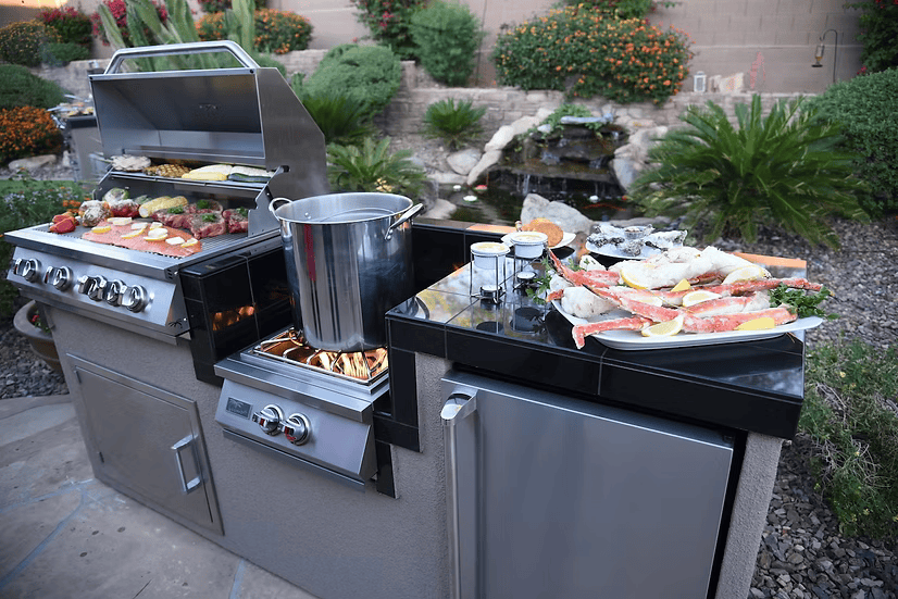 An outdoor kitchen with the Kokomo Grills Power Burner 4 Burner 32-Inch Built-In Barbeque Grill Island Kitchen Package cooks meats, vegetables, and seafood. Sauces and lemon slices sit nearby in a landscaped backyard with lush plants and a small waterfall.