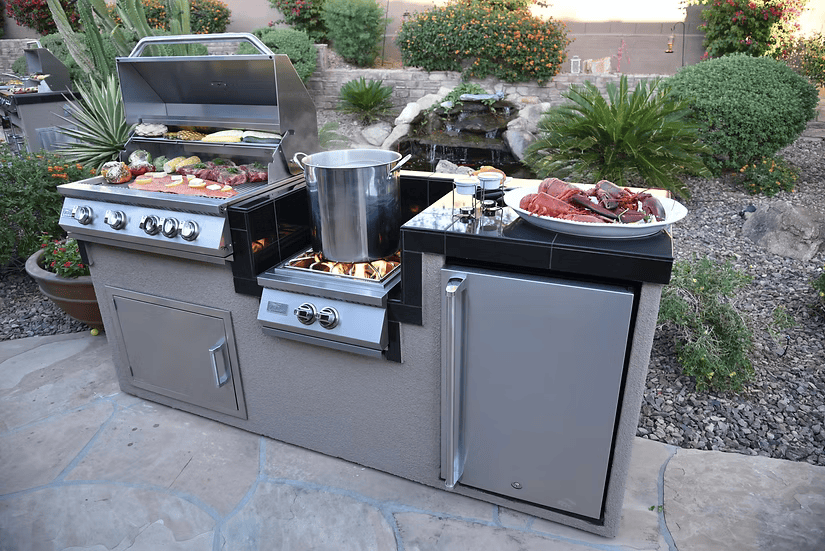 This outdoor kitchen showcases the Kokomo Grills Power Burner 4 Burner 32-Inch Built-In Barbeque Grill Island Kitchen Package, with sizzling food, a burner with a large pot, cooked lobster, plus a mini fridge and cabinets beneath the counter.
