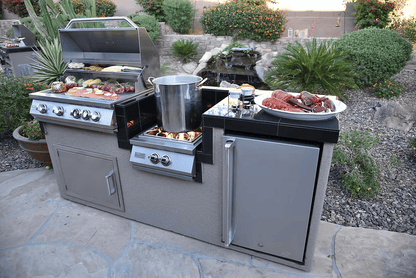 This outdoor kitchen showcases the Kokomo Grills Power Burner 4 Burner 32-Inch Built-In Barbeque Grill Island Kitchen Package, with sizzling food, a burner with a large pot, cooked lobster, plus a mini fridge and cabinets beneath the counter.