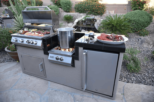 This outdoor kitchen showcases the Kokomo Grills Power Burner 4 Burner 32-Inch Built-In Barbeque Grill Island Kitchen Package, with sizzling food, a burner with a large pot, cooked lobster, plus a mini fridge and cabinets beneath the counter.