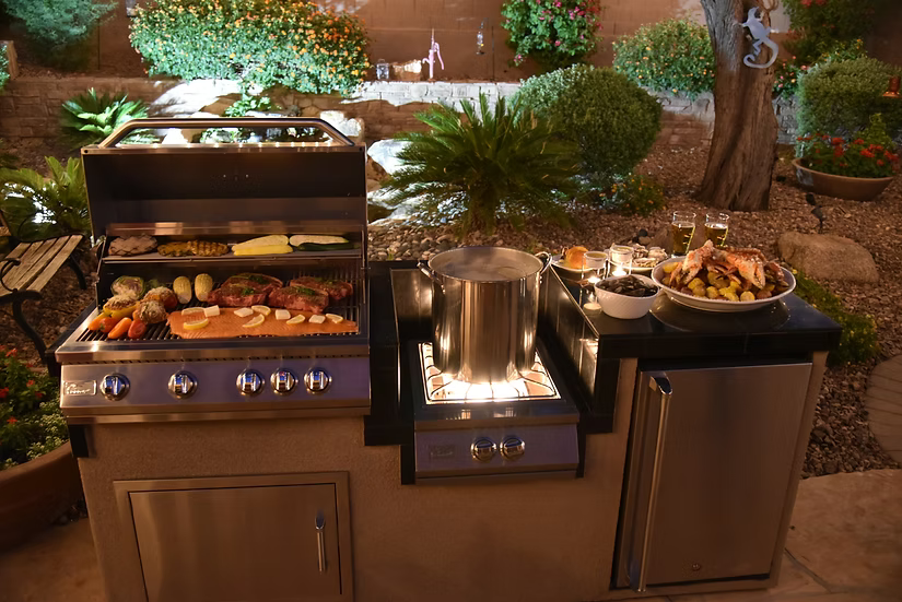 At night, an outdoor kitchen with a Kokomo Grills Professional Built-In Power Burner with LED Lights and a removable wok grate grills meats and veggies, simmers a large pot, and displays food platters against a garden and stone wall backdrop.