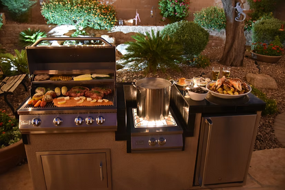 At night, an outdoor kitchen with a Kokomo Grills Professional Built-In Power Burner with LED Lights and a removable wok grate grills meats and veggies, simmers a large pot, and displays food platters against a garden and stone wall backdrop.