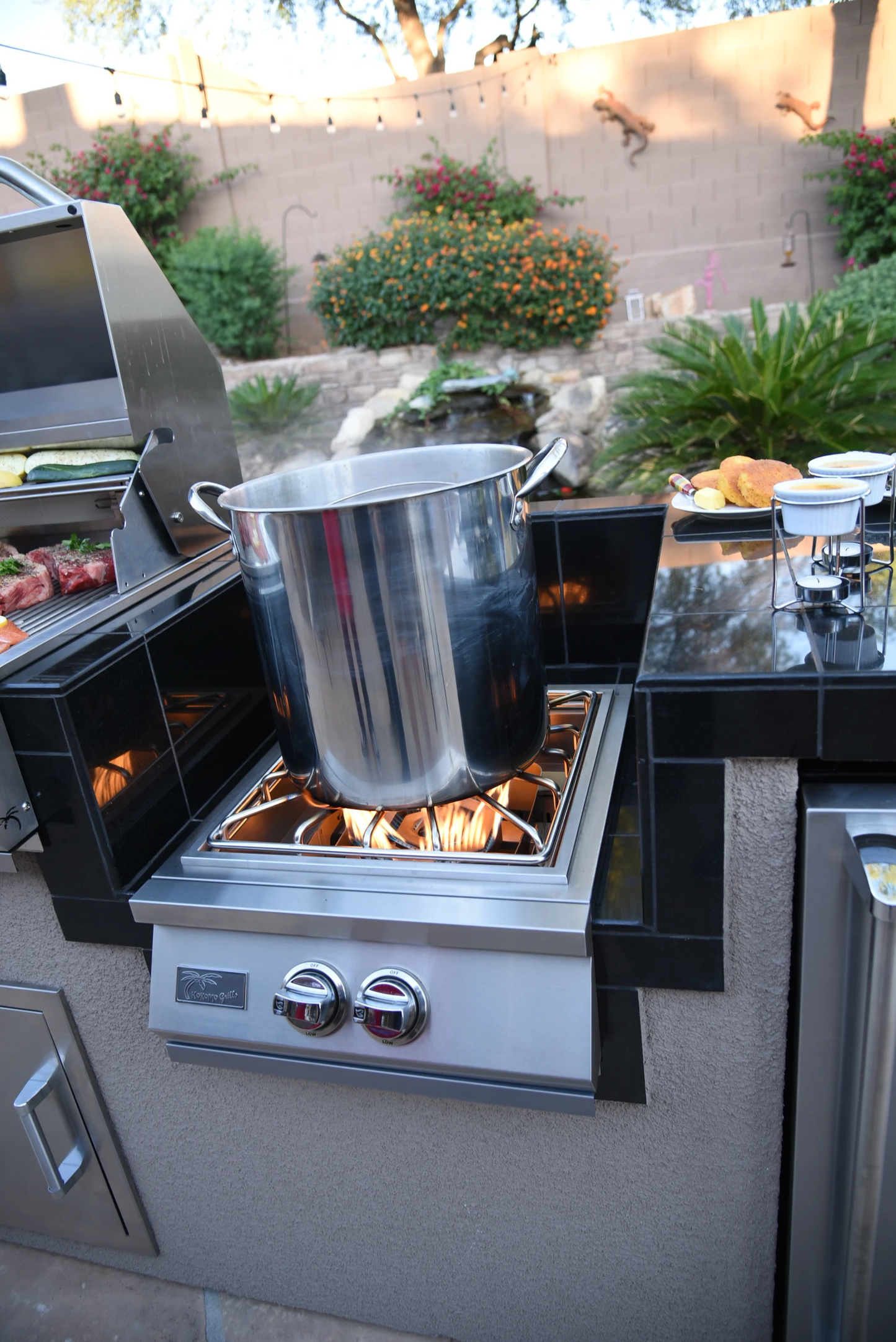 A Kokomo Grills Professional Built-In Power Burner with LED Lights and a removable wok grate sits in an outdoor kitchen with black tiles, a grill, and food on the counter nearby in a backyard setting.