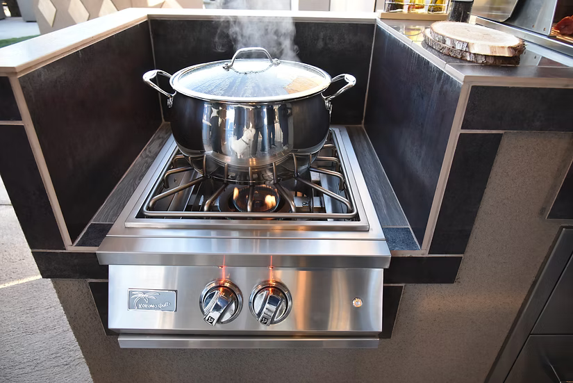 A stainless steel pot with lid steams atop the Kokomo Grills Professional Built-In Power Burner with LED Lights and a removable grate for wok, set in an outdoor kitchen with dark tile and countertops.