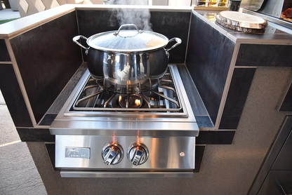 A stainless steel pot with lid steams atop the Kokomo Grills Professional Built-In Power Burner with LED Lights and a removable grate for wok, set in an outdoor kitchen with dark tile and countertops.
