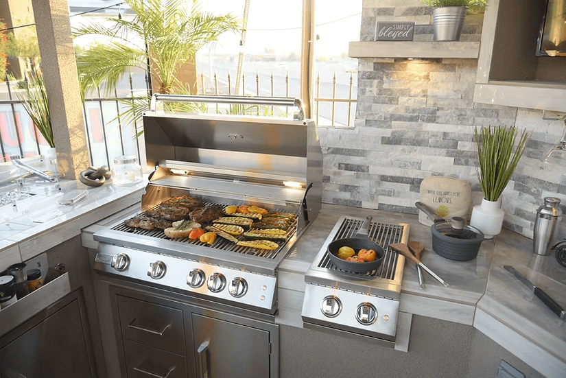 Sunny patio outdoor kitchen featuring a stainless steel grill for meat and veggies, potted plants, modern stone backsplash, and a Kokomo Grills Professional Double Side Burner with Removable Cover for your pot and pan cooking.
