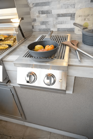 A KoKoMo Grills Professional Double Side Burner with Removable Cover by Kokomo Grills is built into the outdoor kitchen counter, holding a black cast iron pan with tomatoes and bell peppers beside wooden utensils. Grilled food sits on a nearby grill.