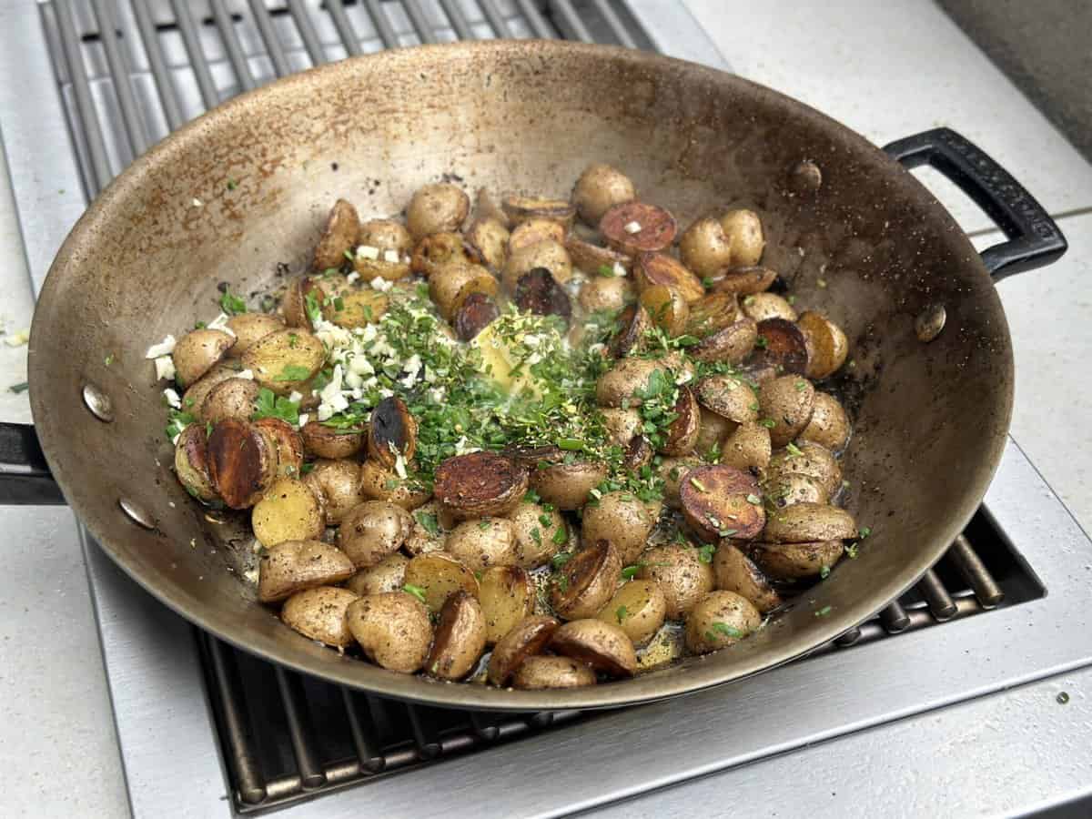 Sliced baby potatoes sauté in a large pan on the KoKoMo Grills Professional Double Side Burner by Kokomo Grills, seasoned with herbs and minced garlic. Fresh green herbs garnish the golden brown potatoes as steam rises from the hot pan.