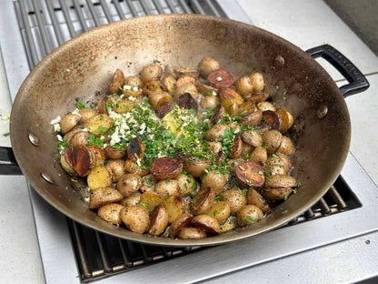 Sliced baby potatoes sauté in a large pan on the KoKoMo Grills Professional Double Side Burner by Kokomo Grills, seasoned with herbs and minced garlic. Fresh green herbs garnish the golden brown potatoes as steam rises from the hot pan.