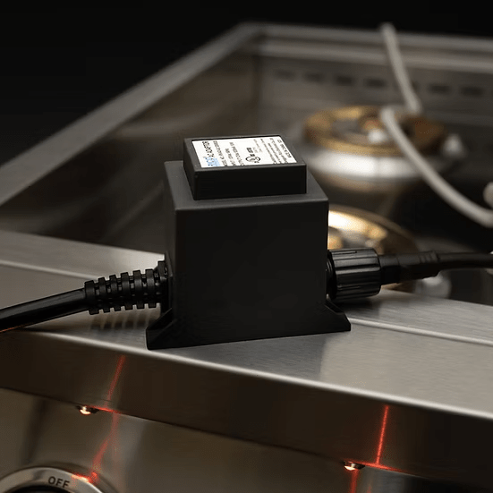 A black device with a label is plugged in and rests on the edge of a KoKoMo Grills Professional Double Side Burner with Removable Cover by Kokomo Grills, featuring visible burners and control knobs in a stainless steel outdoor kitchen setup.