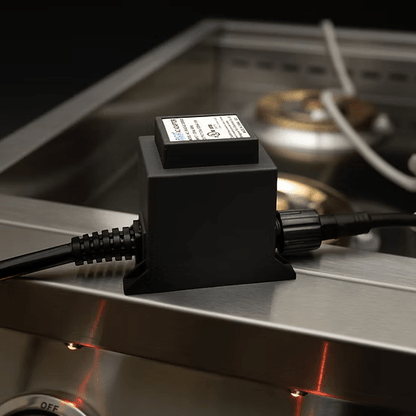 A black device with a label is plugged in and rests on the edge of a KoKoMo Grills Professional Double Side Burner with Removable Cover by Kokomo Grills, featuring visible burners and control knobs in a stainless steel outdoor kitchen setup.