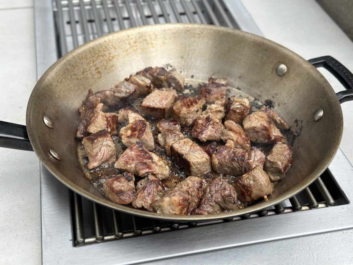 Chunks of beef are searing in a large skillet on a stove, their browned surfaces and juices visible as they cook over high heat on the KoKoMo Grills Professional Double Side Burner with Removable Cover by Kokomo Grills.