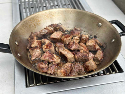 Chunks of beef are searing in a large skillet on a stove, their browned surfaces and juices visible as they cook over high heat on the KoKoMo Grills Professional Double Side Burner with Removable Cover by Kokomo Grills.