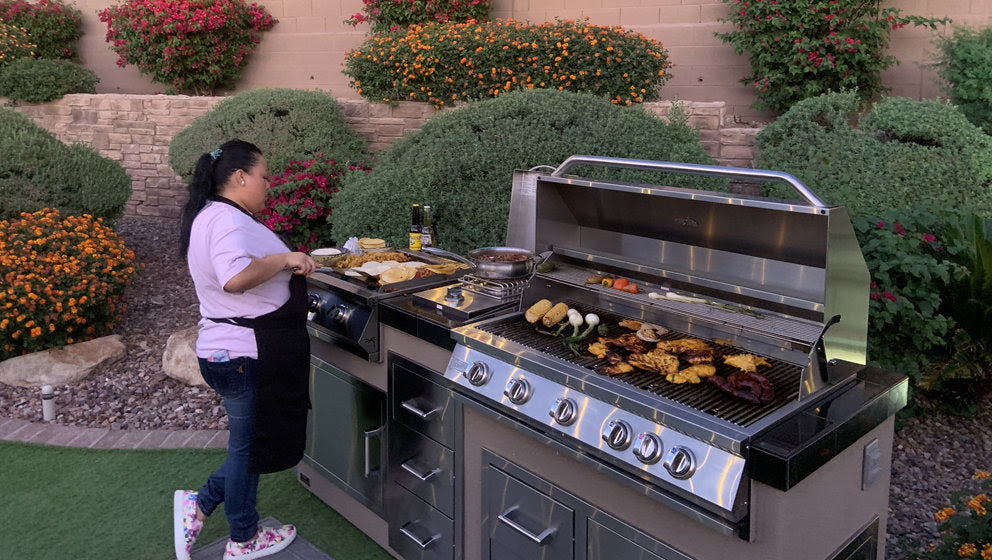 A woman cooks meats and vegetables on the Kokomo Grills 40-Inch Built-In Gas Grill (5 Burner / Back Burner) by Kokomo Grills in her garden, surrounded by flowering bushes, wearing a pink shirt, apron, and sneakers in her outdoor kitchen.