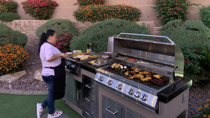 A woman cooks meats and vegetables on the Kokomo Grills 40-Inch Built-In Gas Grill (5 Burner / Back Burner) by Kokomo Grills in her garden, surrounded by flowering bushes, wearing a pink shirt, apron, and sneakers in her outdoor kitchen.