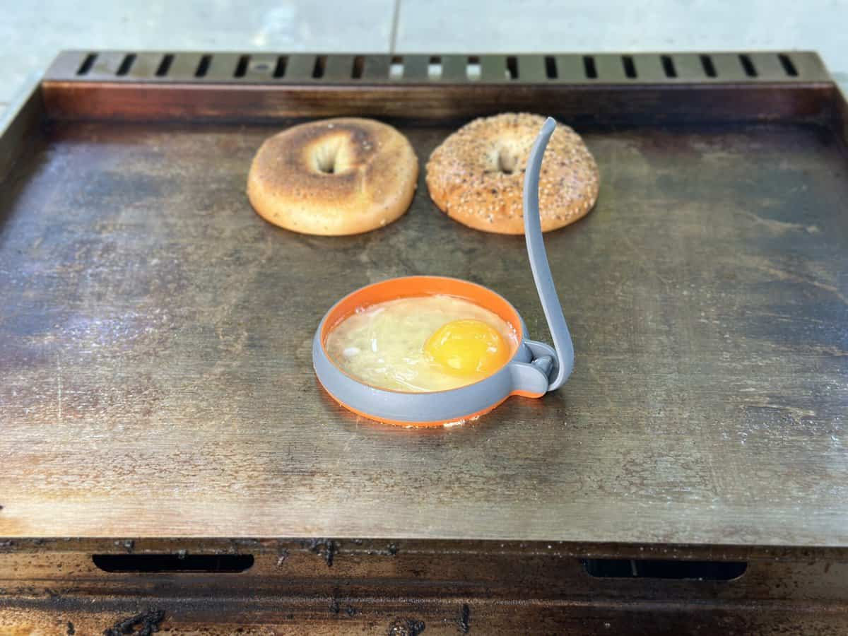 Two bagel halves toast on the Kokomo Grills Professional Griddle 30-Inch Outdoor Kitchen Teppanyaki Flat Grill, while a raw egg cooks in an orange silicone ring. The griddle's 304 stainless steel surface looks richly seasoned.