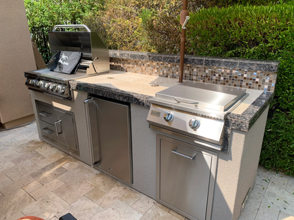 Outdoor kitchen setup with the Kokomo Grills Professional Griddle 30-Inch Teppanyaki Flat Grill, side burner, mini fridge, and storage cabinets on a stone countertop with tile backsplash, surrounded by lush greenery.