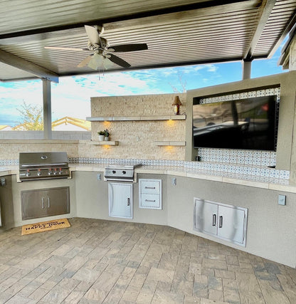 Outdoor kitchen with a Kokomo Grills Professional Griddle 30-Inch Teppanyaki Flat Grill, stove, storage cabinets under a covered patio. Wall shelves, large TV, accent lighting, tiled countertops, and paver flooring create a modern setup.