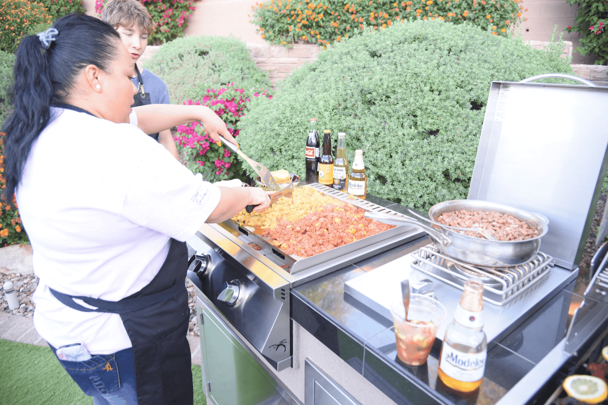 A woman prepares a feast of meat and veggies on the Kokomo Grills Professional Griddle 30-Inch Outdoor Kitchen Teppanyaki Flat Grill, with bottles, drinks, and a pan nearby; blooming bushes and flowers enhance the background.