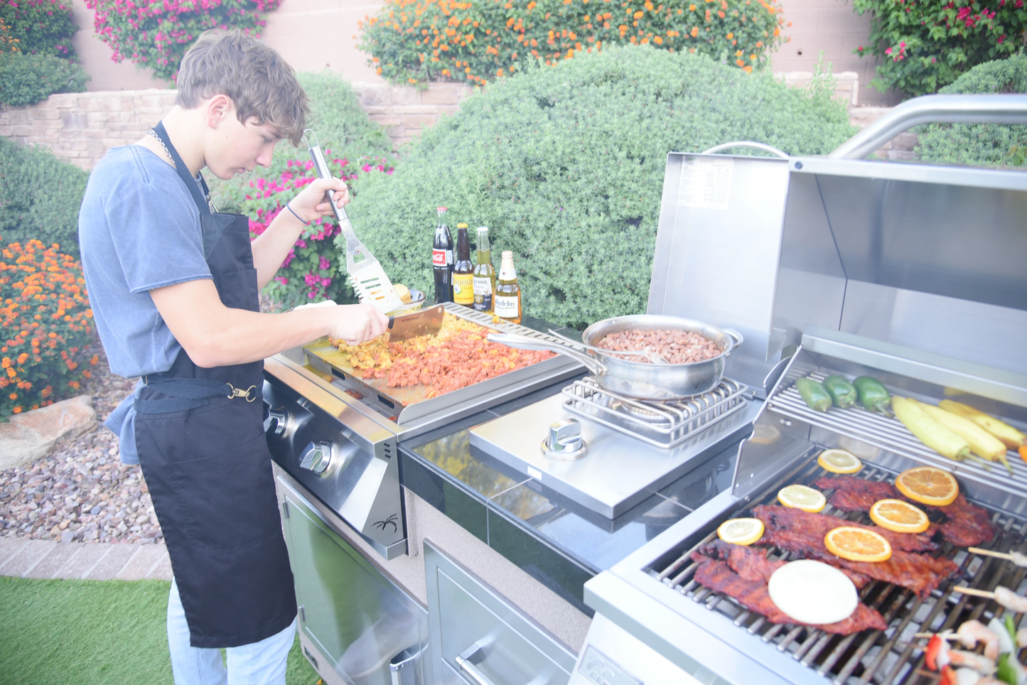 Wearing a black apron, a person cooks ribs, corn, and peppers on the Kokomo Grills Professional Griddle 30-Inch Outdoor Kitchen Teppanyaki Flat Grill by Kokomo Grills, with sauces and beers nearby amid lush greenery and vibrant orange flowers.