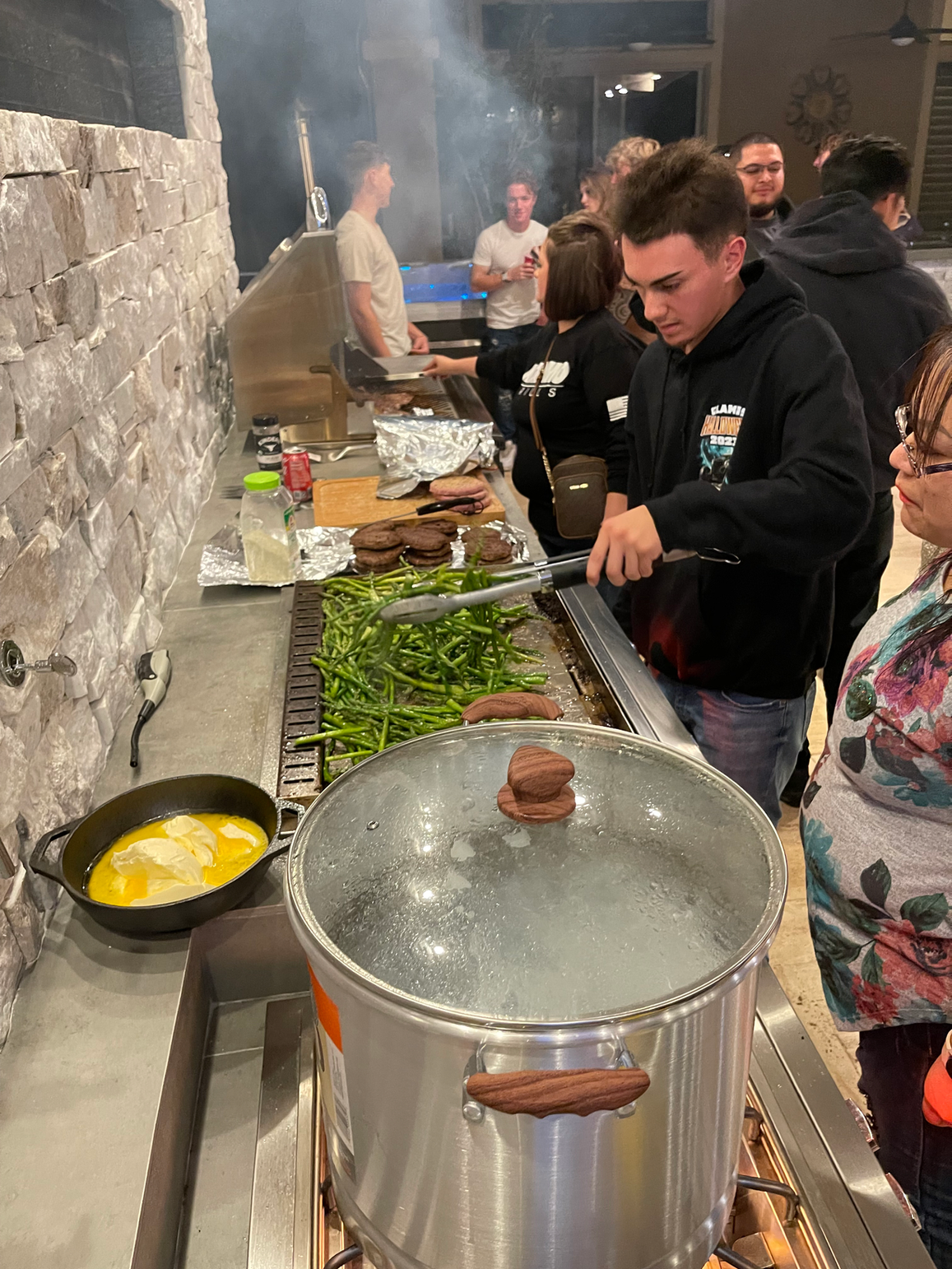 Friends gather around the Kokomo Grills Professional Griddle 30-Inch Outdoor Kitchen Teppanyaki Flat Grill as green beans and burger patties sizzle. A steaming pot sits by the 304 stainless steel grill, with a buttered skillet nearby and smoke rising in the background.