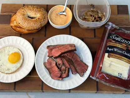 A sesame bagel, sliced steak, and sunny-side-up egg, topped with caramelized onions, sauce, and white cheddar—perfectly prepared on the Kokomo Grills Professional Griddle 30-Inch Outdoor Kitchen Teppanyaki Flat Grill.