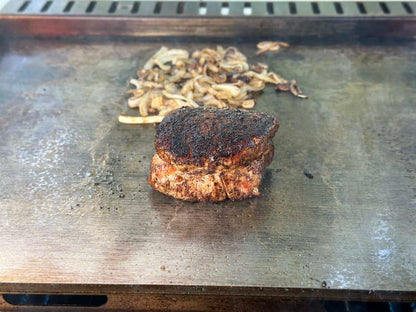 A sizzling steak cooks on the Kokomo Grills Professional Griddle 30-Inch Outdoor Kitchen Teppanyaki Flat Grill, with sautéed onions and mushrooms nearby. The 304 stainless steel surface displays perfect browning and beautiful grill marks.