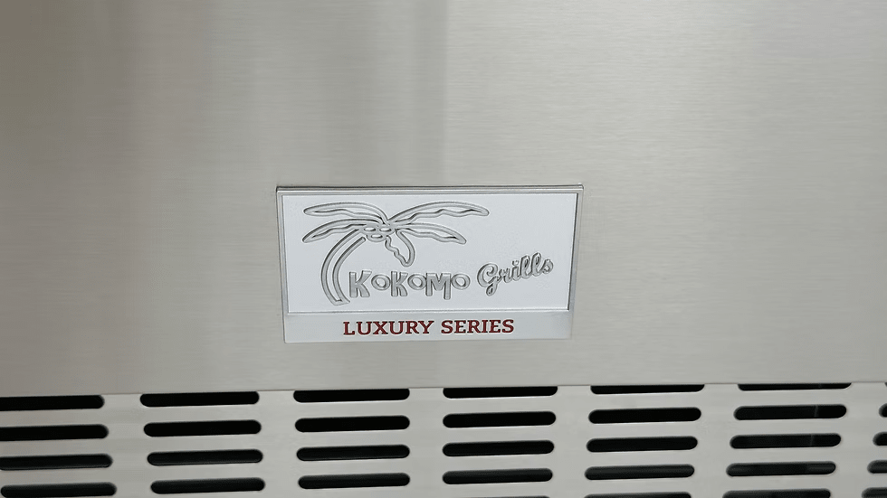 A metal plate on the Kokomo Grills Professional Luxury Outdoor Kitchen Kegerator KO-LUX-KEG features a palm tree graphic and “Kokomo Grills” above a vented grill, ideal for your kegerator or outdoor kitchen setup.