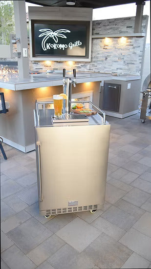 The Kokomo Grills Professional Luxury Outdoor Kitchen Kegerator KO-LUX-KEG, featuring a dual tap system with two filled glasses, sits on a patio near a modern grill station with a Kokomo Grills sign, stone backsplash, and outdoor lighting.