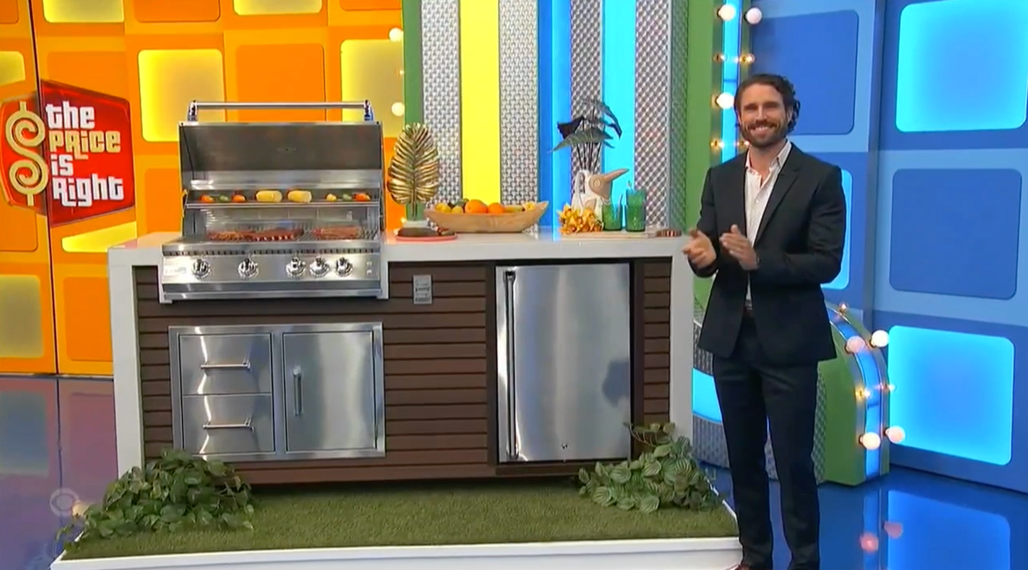 A smiling man in a suit stands beside the Kokomo Grills Professional Shiplap Built-In BBQ Island Outdoor Kitchen with Waterfall Edge, topped with fruit and drinks. The Price Is Right logo appears in the colorful background on the left.