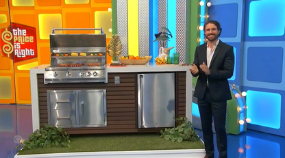 A smiling man in a suit stands beside the Kokomo Grills Professional Shiplap Built-In BBQ Island Outdoor Kitchen with Waterfall Edge, topped with fruit and drinks. The Price Is Right logo appears in the colorful background on the left.