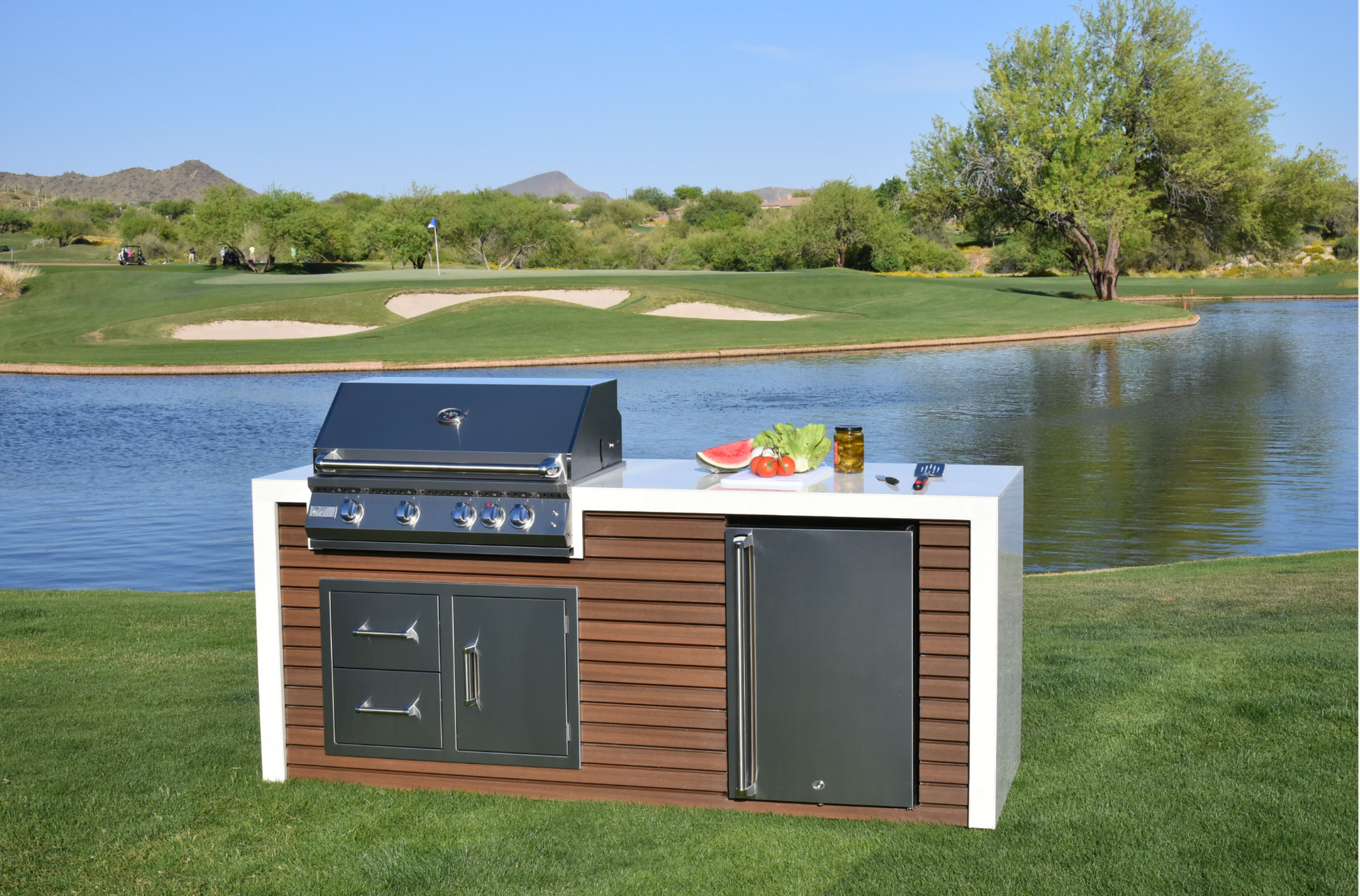 A Kokomo Grills Professional Shiplap Built-In BBQ Island Outdoor Kitchen with Waterfall Edge sits on a green lawn by a pond, golf course, and mountains. Fresh vegetables and watermelon slices are displayed on the counter.