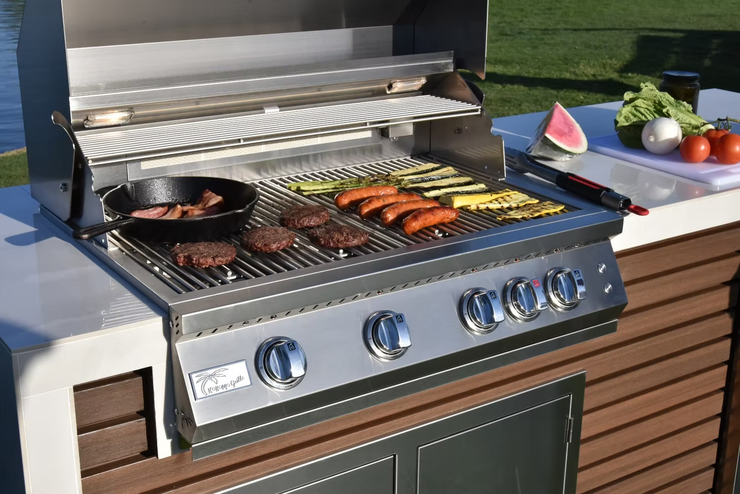 The Kokomo Grills Professional Shiplap Built-In BBQ Island Outdoor Kitchen with Waterfall Edge by Kokomo Grills sizzles with burgers, sausages, bacon in a skillet, and zucchini while fresh produce sits on the counter—all in a sunny yard.