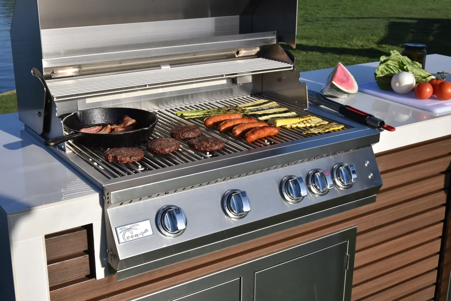 The Kokomo Grills Professional Shiplap Built-In BBQ Island Outdoor Kitchen with Waterfall Edge by Kokomo Grills sizzles with burgers, sausages, bacon in a skillet, and zucchini while fresh produce sits on the counter—all in a sunny yard.