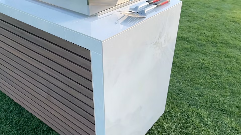 A close-up of the Kokomo Grills Professional Shiplap Built-In BBQ Island Outdoor Kitchen with Waterfall Edge on a lawn. Three stainless steel barbecue tools with red and black handles rest on its white countertop.