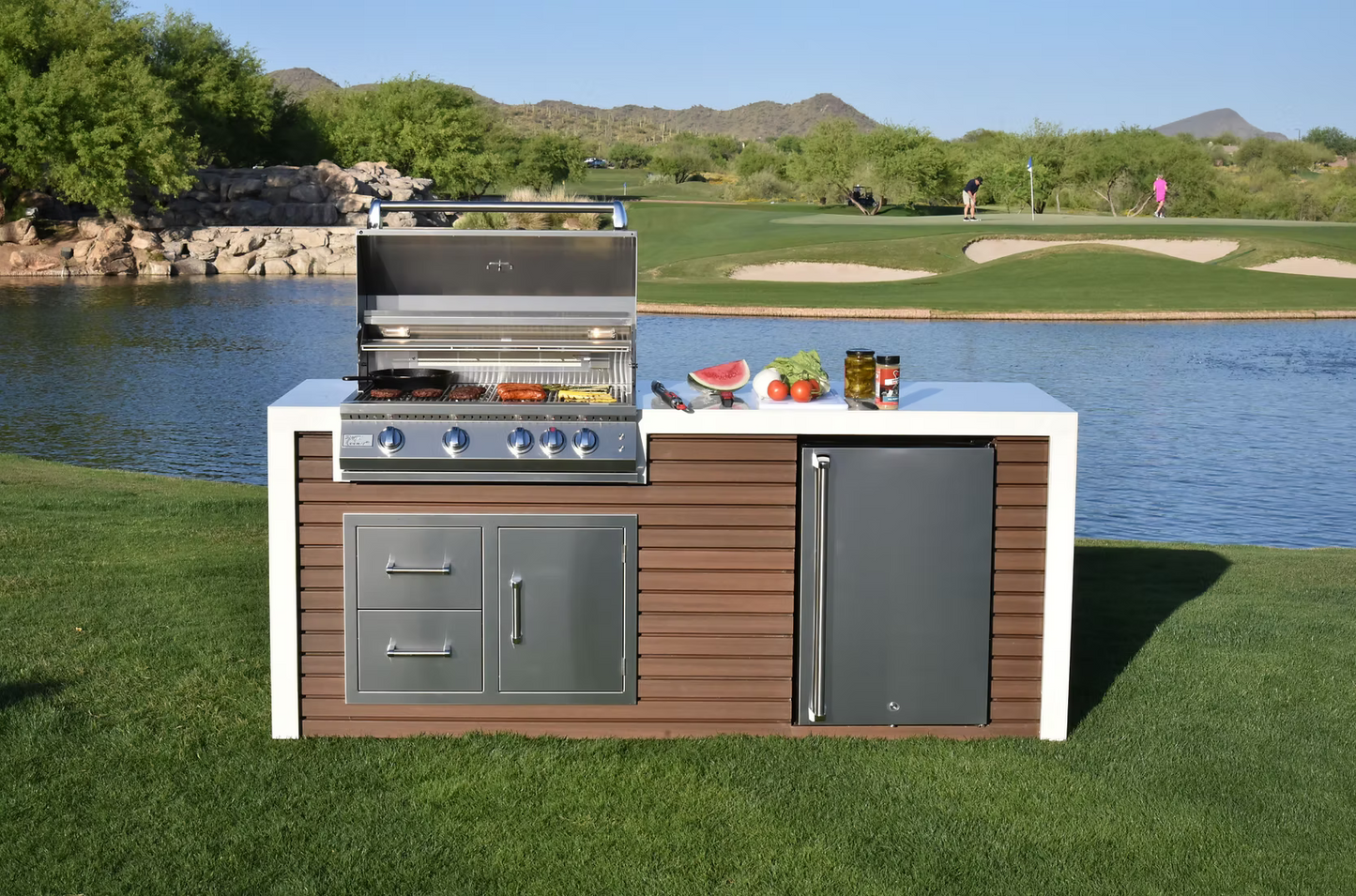 The Kokomo Grills Professional Shiplap Built-In BBQ Island Outdoor Kitchen with Waterfall Edge by Kokomo Grills features a grill, stove, and refrigerator on grass beside a lake. Fresh produce sits on the counter; golfers and mountains appear in the clear-sky background.