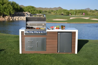 The Kokomo Grills Professional Shiplap Built-In BBQ Island Outdoor Kitchen with Waterfall Edge by Kokomo Grills features a grill, stove, and refrigerator on grass beside a lake. Fresh produce sits on the counter; golfers and mountains appear in the clear-sky background.