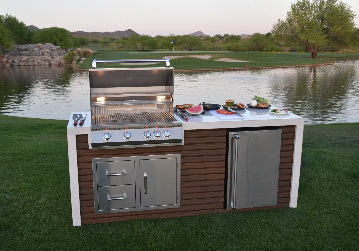 Lakeside outdoor kitchen featuring the Kokomo Grills Professional Shiplap Built-In BBQ Island with Waterfall Edge, stainless steel refrigerator, plates of food, and grilling utensils on grass with trees and hills in the background.