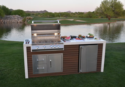 Lakeside outdoor kitchen featuring the Kokomo Grills Professional Shiplap Built-In BBQ Island with Waterfall Edge, stainless steel refrigerator, plates of food, and grilling utensils on grass with trees and hills in the background.
