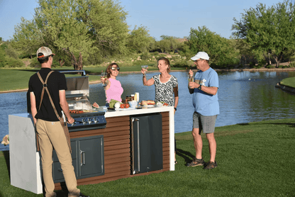 Four adults enjoy drinks and laughter by a Kokomo Grills Professional Shiplap Built-In BBQ Island Outdoor Kitchen with Waterfall Edge, surrounded by green grass and trees for the perfect outdoor gathering.