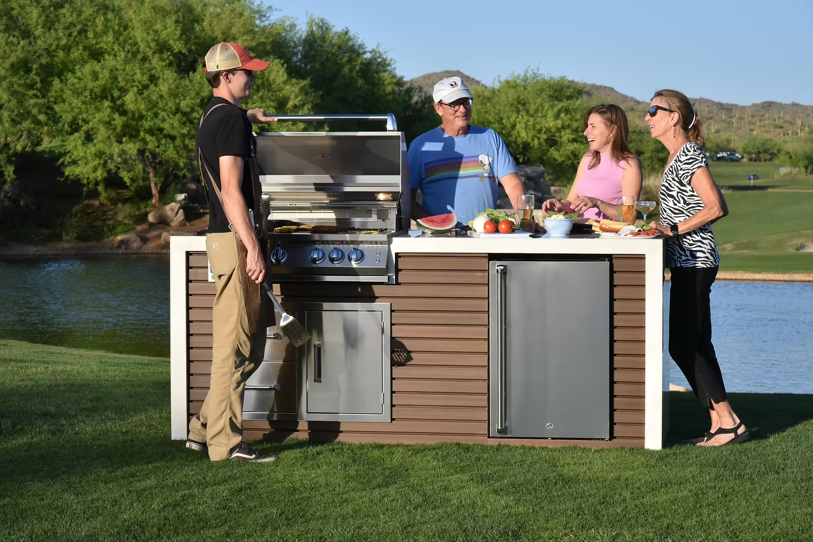 Four people gather by a Kokomo Grills Professional Shiplap Built-In BBQ Island Outdoor Kitchen with Waterfall Edge, smiling and chatting as food cooks. Watermelon and fresh ingredients rest on the countertop against a scenic lakeside backdrop.