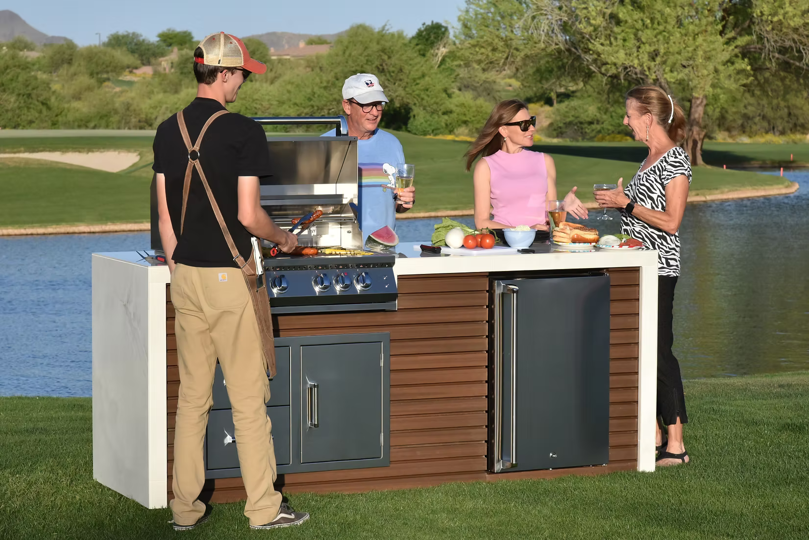 Four adults gather by a Kokomo Grills Professional Shiplap Built-In BBQ Island Outdoor Kitchen with Waterfall Edge, smiling and chatting as they prepare food and drinks together on a sunny lakeside day.