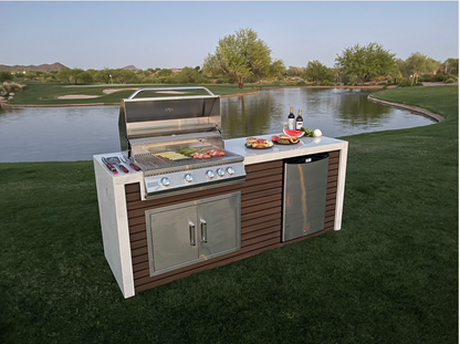 A Kokomo Grills Professional Shiplap Built-In BBQ Island Outdoor Kitchen with Waterfall Edge stands on lush grass near a pond, food sizzling on the grill, wine bottles and plates arranged atop, framed by trees and hills under a clear sky.